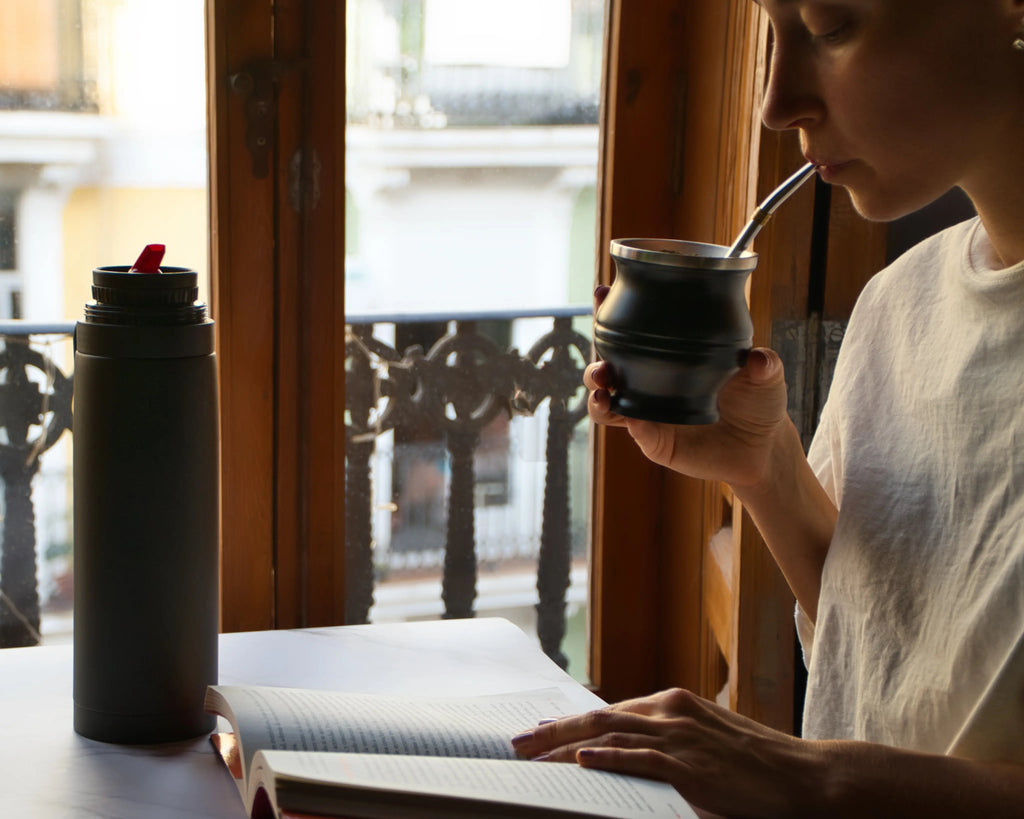 Persona toma mate junto a la ventana, con un libro abierto y un termo sobre la mesa