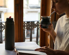 Persona toma mate junto a la ventana, con un libro abierto y un termo sobre la mesa